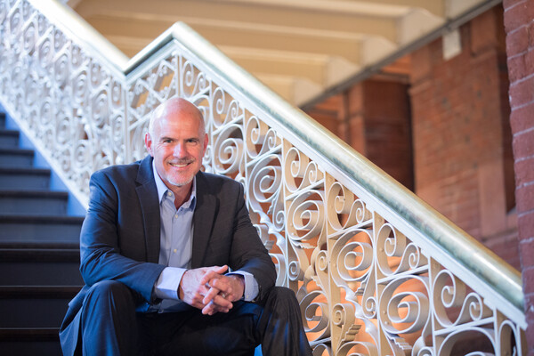 A person in a suit and button-down shirt sitting on a stairwell landing, smiling. The intricate white stairwell and a brick wall behind it are to the person's right.