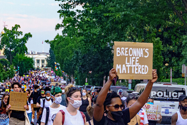 Large crowd wearing masks protesting in the streets of D.C., person in foreground holds a sign reading BREONNA’S LIFE MATTERED.