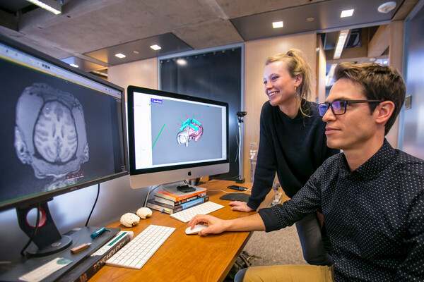 Two people in front of two computer monitors. The person moving the mouse is sitting. The other person stands watching. 