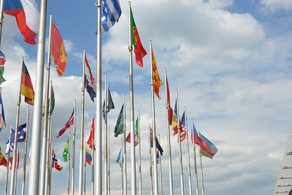 Flags of various nations wave atop flagpoles against a blue sky with clouds