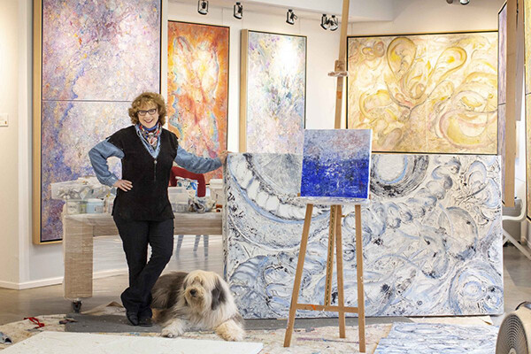 Jill Krutick standing in her studio surrounded by paintings with a fluffy dog at her feet.