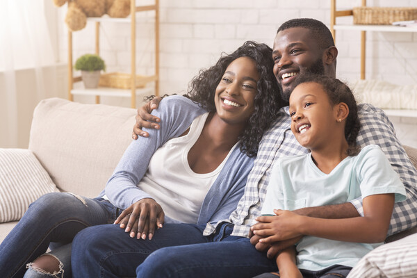 Sitting on a couch embracing and smiling is a Black mom, dad, and daughter.