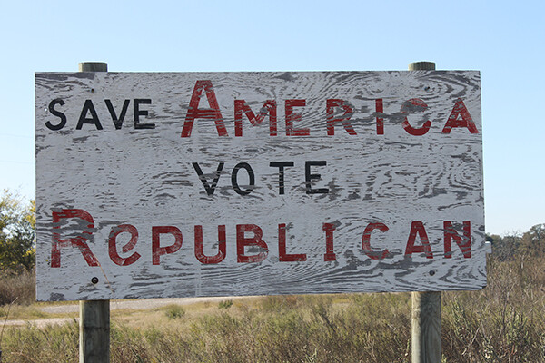 A faded wooden sign reading "save America vote Republican" stands amid scrubby plants on a Texas roadside.