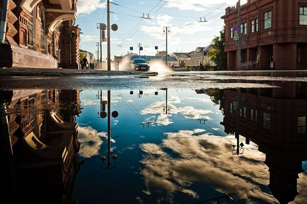 Reflection of a blue sky in a large puddle on a city street with a car driving through wet streets in the sunshine after a rain shower.