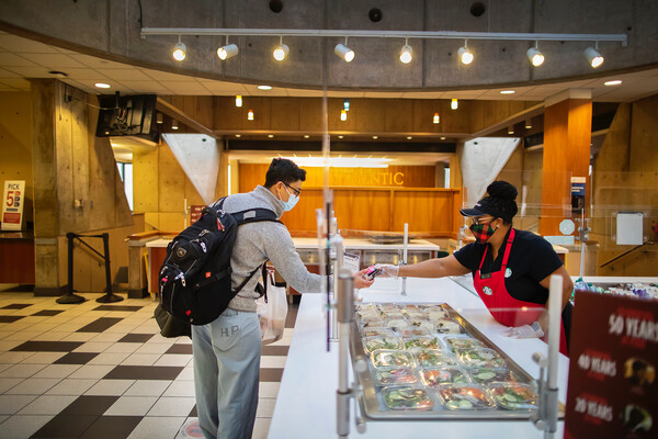 Cafeteria staff person wearing mask and apron handing food to a student on the other side of the counter who is also wearing a mask with a backpack on 