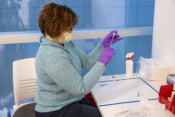 A person at a desk wearing rubber gloves and a mask puts a dose of medication into a syringe.