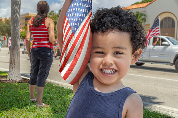 A young child smiles at the camera standing in green grass holding a small American flag next to his head as a pickup truck passes on the street behind him with a passenger waving an American flag.