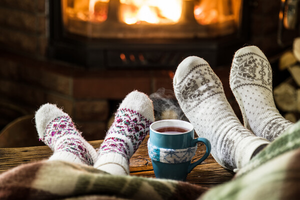 Two pairs of socked feet, up on a bench next to a steaming mug of hot liquid. In the background is a fireplace with a fire.