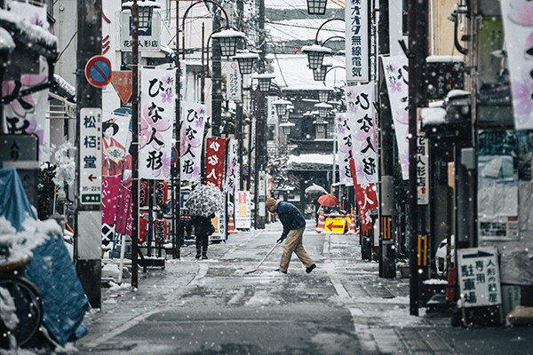Man shovels snowy street, which is lined by lanterns and banners with Japanese characters