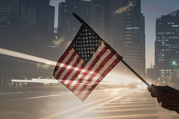 A hand is seen waving a small American flag as traffic zips by on a city street at dusk