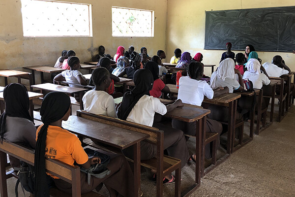 Classroom full of girls seated at wooden desks with Fatima Al Rashed seated in front of the blackboard in the front of the class.