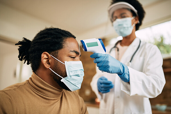 African American person wearing a face mask having their temperature taken via forehead scanner by a masked, gloved medical professional.