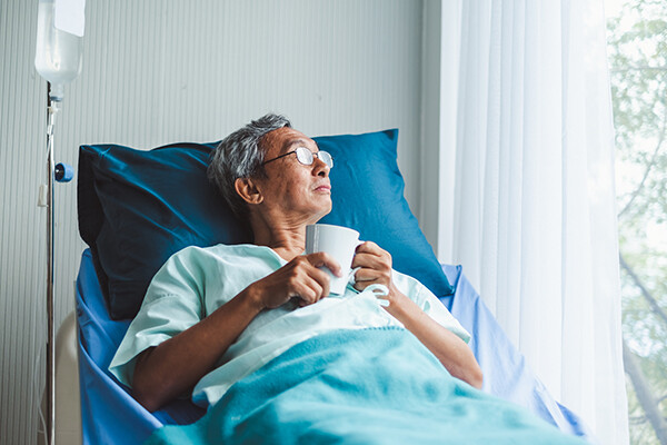 Recovering hospital patient sitting up in hospital bed holding a cup of tea looking out the window.