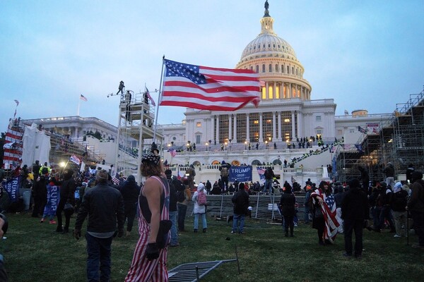 A person in an American flag outfit and bandana carrying an American flag in front of a large group of people at the U.S. Capitol building. Many hold flags that read "Trump 2020."