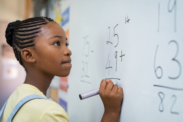 A young girl doing a math problem at a whiteboard. The numbers 25, 49, and 14 are visible.