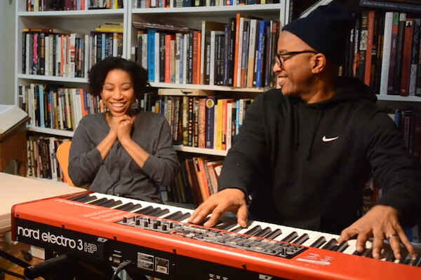 Father plays keyboard while his daughter smiles. Bookshelves in background. 