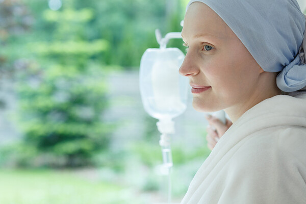 Cancer patient with IV drip and scarf on their hair looks out the window.