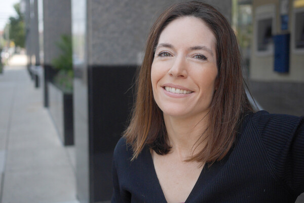 Close-up of a smiling person in a black V-neck shirt, standing outside near marble pillars.