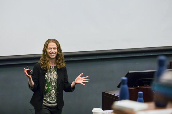 A person standing in the front of a classroom holding a pointer, smiling. Blurry water bottles appear in the foreground. 