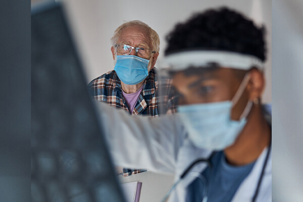 A masked doctor examines brain scans of an elderly masked patient seated in the background.