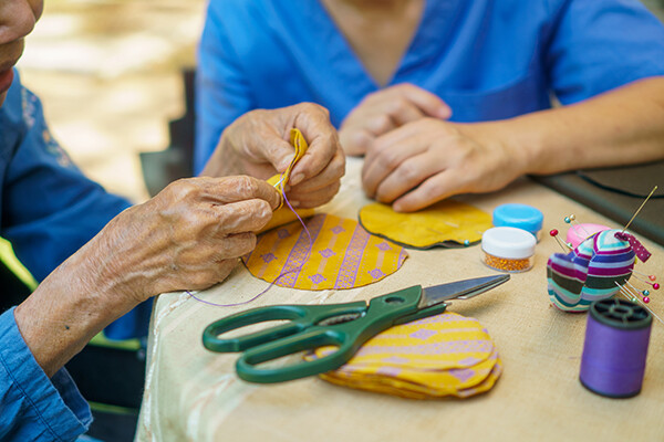Elderly hands sewing with a needle and thread at a table with sewing supplies, a younger pair of hands assists at the side.
