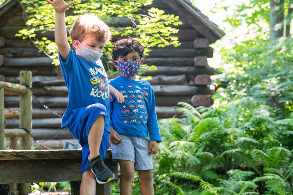 two children wearing masks in front of a log cabin and lush ferns