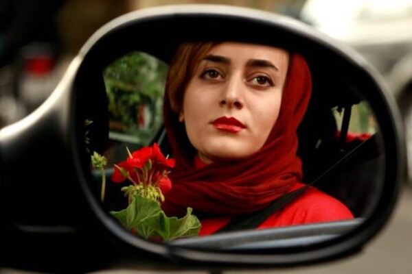 A woman with a red headscarf, red red jacket and red lipstick holding a red geranium is seen in a reflection in a car's side mirror