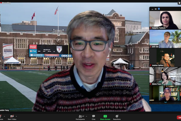 Person wearing glasses speaks on a Zoom call in front of a background featuring Penn's football stadium, as five others on the call are in a vertical column on the right side of the screen