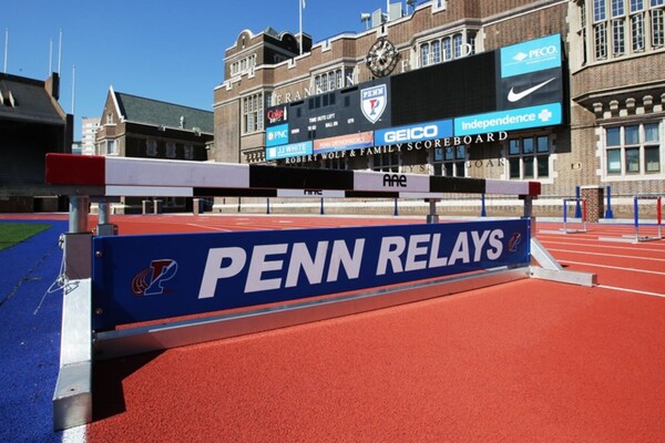 On the track around Franklin Field, a Penn Relays banner rests on the bottom of a hurdle bar.