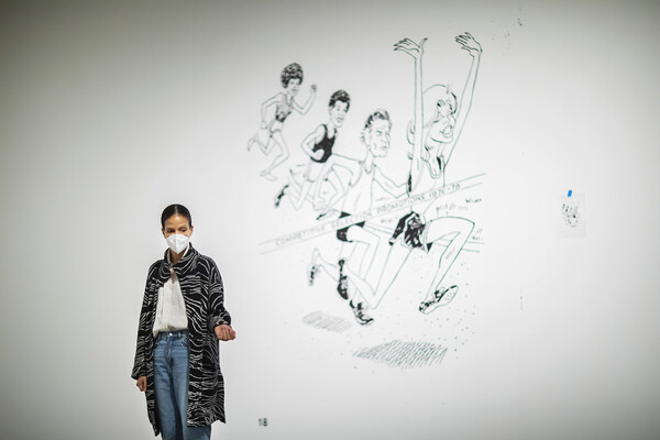 Artist standing in gallery in front of an illustration painted on the wall of people running a race 