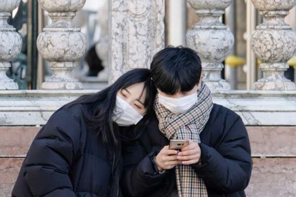 Two people in black jackets wearing white face masks sit in front of marble columns, with the person on the left putting her head on the other person's shoulder, who is looking at a smartphone.