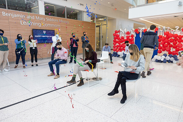 Perelman School of Medicine students sit masked and socially distanced surrounded by balloons and streamers and confetti as they open envelopes.