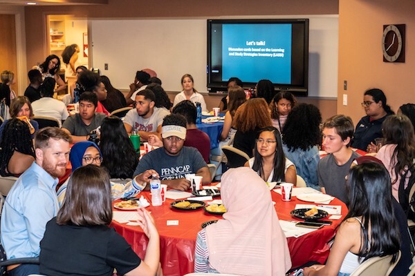 Students and staff sitting and eating at round tables. A screen in the back reads, "let's talk!"