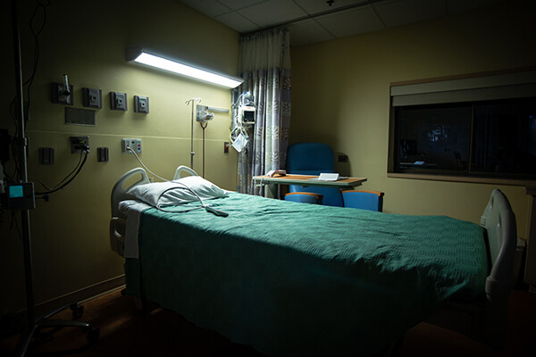 A hospital room with all lights off but the fluorescent light above bed, which has a green blanket. Behind the bed is a hospital tray that swivels and a blue chair. On the yellow wall behind are about 8 outlets.