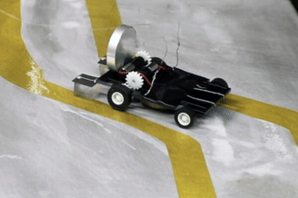 Film still of a small wheeled robot traveling a path between yellow tape.