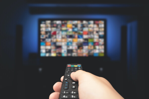 A hand holding a television remote, pointed at a blurry TV straight ahead. On the TV are many colored boxes signifying many show options.