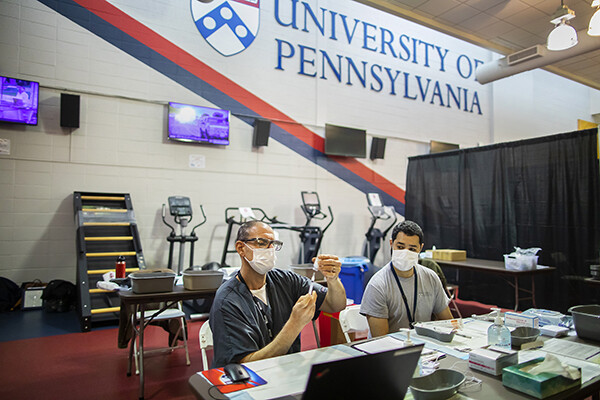 Two people wearing face masks seated at a table, one is preparing a vial of vacccine with a syringe, a University of Pennsylvania shield and logo is on the back wall.