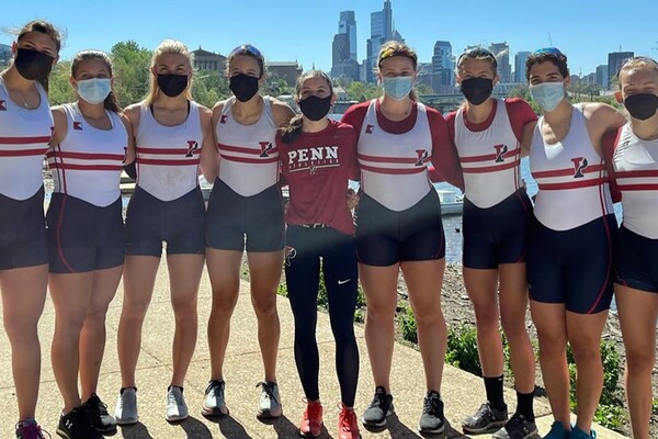 The women's rowing team, wearing masks, poses arm in arm after winning the Kelly Cup on the Schuylkill River.