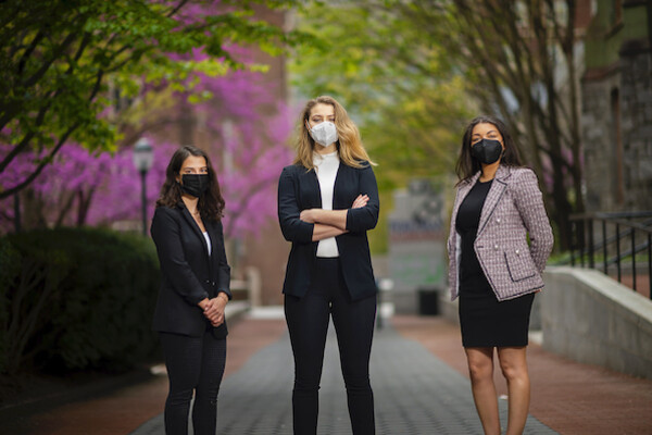 Three women wearing face masks and office dress clothes stand on a pathway with a tree full of pink flowers behind them