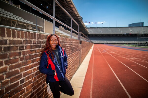 Uchechi Nwogwugwu, wearing a red and blue Penn jacket, leans against a brick wall at Franklin Field, next to the track.