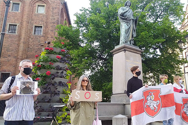 Six people stand in a square in Poland in front of a statue of a man in toga, some hold Belarusian flags, one woman holds a sign reading "SOS" and a man holds a photo of Belarusian journalist Raman Pratasevich
