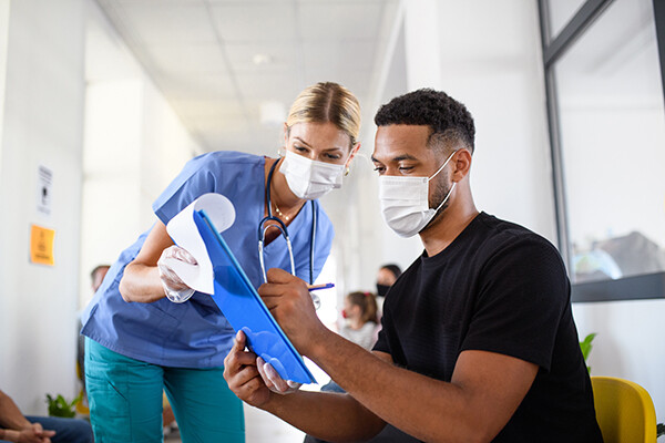 Medical worker in a face mask holds a clipboard for an African American person to sign.