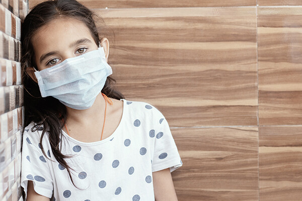 A young person wearing a mask and polka dot t-shirt leaning against a faux wooden wall.