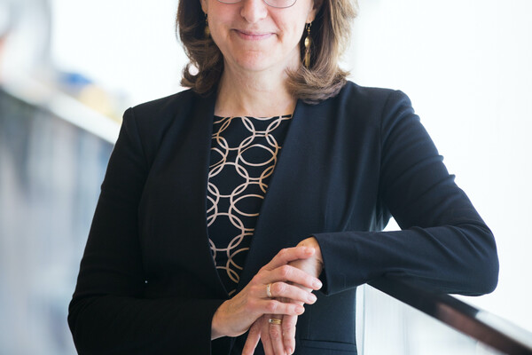 A person leaning against a glass railing, one elbow on the rail, the other hand on top of the leaning hand. The person is wearing a black dress with gold intertwining circles, a black sweater, glasses and gold earrings.