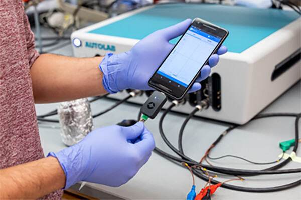 Gloved hands holding a smart phone with a cord plugged in that is attached to lab equipment.