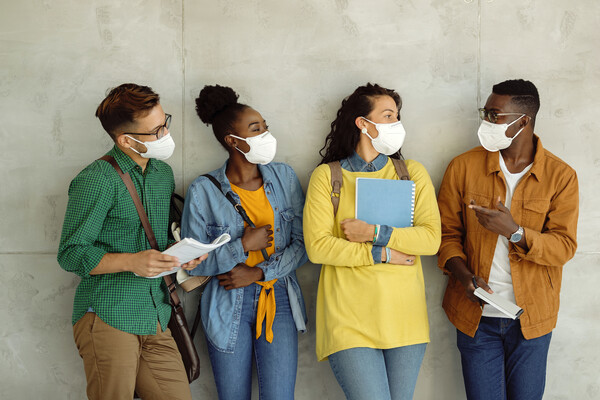 A group of four people leaning against a wall. The one farthest on the left is wearing a bag diagonal across the chest and holding papers. The second from left has on a purse. The second from right has on a backpack and is holding a blue spiral notebook. The person all the way on the right is pointing to the others and holds a folded piece of white paper and a writing utensil. 