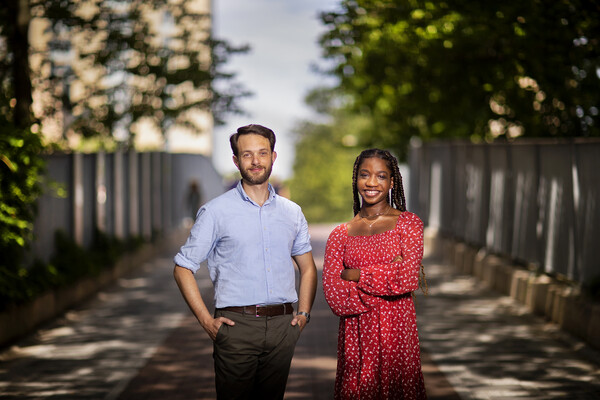 Two people standing outside, with a bridge and trees blurry in the background. One, in a blue button-down shirt and khakis, stands with hands in pockets. The other, in a red dress, stands with arms crossed.