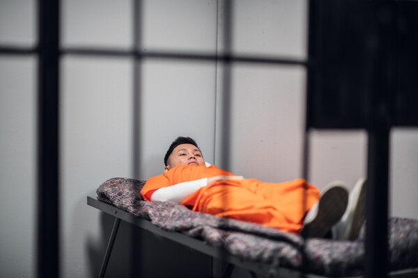 Asian prisoner in jumpsuit laying on a cot behind bars.