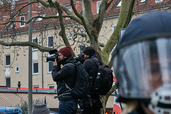 Two journalists take photos in a crowd with a person wearing a face shield in the foreground.