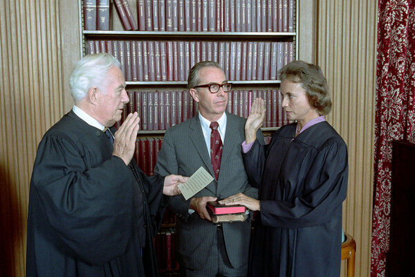 Three people stand in front of a bookcase full of books in burgundy binding, the man on the left is wearing judge robes and has his right hand in the air, the woman on the right is in judge robes and has her right hand in the air and left hand on a bible and a man in the middle wears a suit and tie, is holding the bible and is looking at the woman